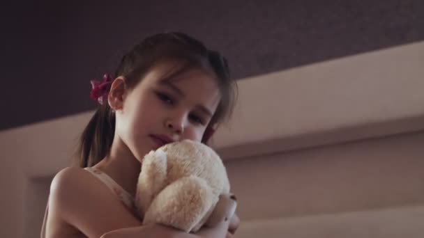 Little girl flowered dress sits on the bed and hugs teddy bear. — Stock Video