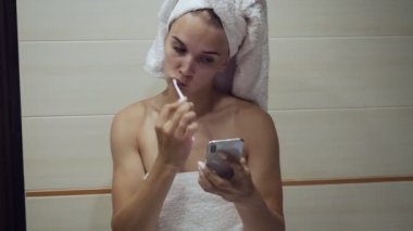 Dişlerini fırçalayan genç kadın akıllı telefona bakıyor.