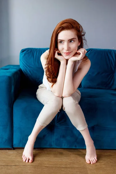 自宅の青いソファに座っている赤毛の女性. — ストック写真