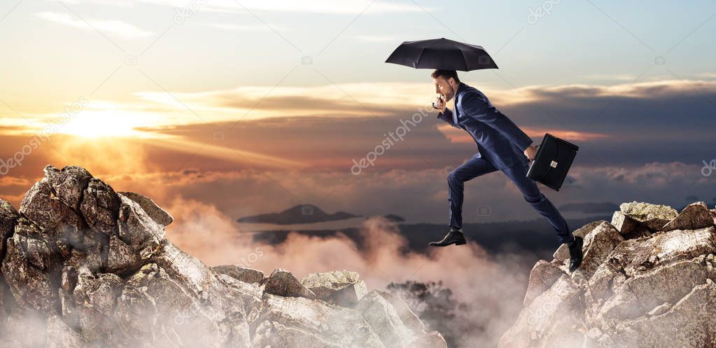 Businessman jumping over a cliff with umbrella and briefcase.