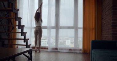 Genç kadın perdeleri açar ve büyük panoramik pencere yakınında dans.