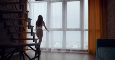 Genç kadın perdeleri açar ve büyük panoramik pencere yakınında dans.