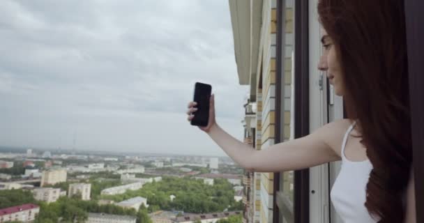 Mujer joven mirando desde la ventana y hace selfie en el teléfono inteligente . — Vídeo de stock