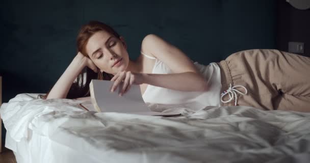 Femme rousse couchée et se détendre sur le lit et regarde dans son journal intime . — Video