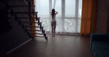 Genç kadın perdeleri açar ve büyük panoramik pencere yakınında dans.
