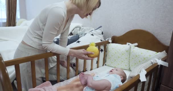 Νεαρή μητέρα στέκεται κοντά στην κούνια δείχνει κίτρινο παιχνίδι στο νεογέννητο μωρό. — Αρχείο Βίντεο
