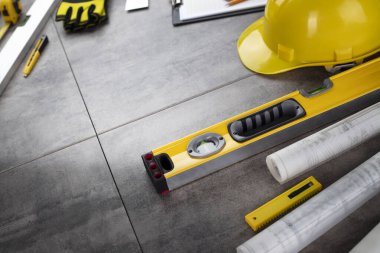 Müteahhit konsepti. Müteahhidin alet çantası, sarı başlık, libella, el testeresi. Gri karoların arka planındaki planlar ve defterler.