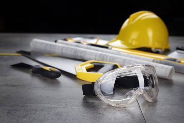 Müteahhit konsepti. Müteahhidin alet çantası, sarı başlık, libella, el testeresi. Gri karoların arka planındaki planlar ve defterler.