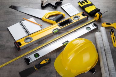 Müteahhit konsepti. Müteahhidin alet çantası, sarı başlık, libella, el testeresi. Gri karoların arka planındaki planlar ve defterler.