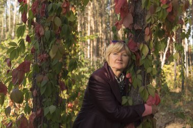 Sonbahar başlarında ormanda mutlu orta yaşlı bir kadın var. Pembe kıyafetler. Güzel ağaçların yakınında duruyor. Etrafında büyük pembe yaprakları vardır.