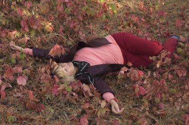 Sonbahar başlarında ormanda mutlu orta yaşlı bir kadın var. Pembe kıyafetler. Çim üzerinde yatıyor. Etrafında büyük pembe yaprakları vardır.