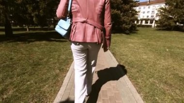 Omzunda çanta olan pembe ceketli bir kadın taştan yapılmış bir yolda yürüyor. Yeşil çimenler ve uzun ladin. Arkadan ve omuz uzalaktan çekim.