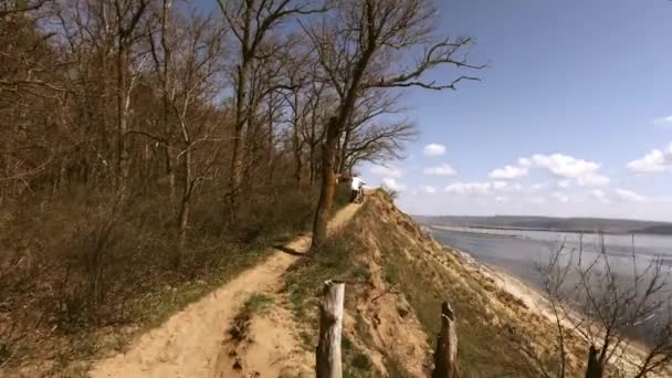 一个戴着红帽子的中年妇女 他沿着森林中的一条小路骑自行车 右高悬崖和大河 晴朗的日子 以速度从背面拍摄 — 图库视频影像
