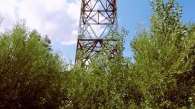 Yüksek voltaj desteği ormanda duruyor. Büyük metal parçaların tasarımı. Kamera hareketi yukarı. Mavi gökyüzü ve bulutlar görmek.