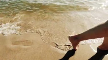 Saf sarı kum ve berrak su küçük dalgalar. Kadın yalınayak gidiyor. Yakın çekim ve sadece bacaklar görünür. Yavaş çekim. Atmosferik video.