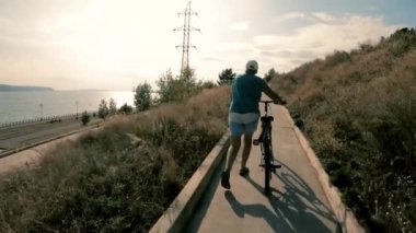 Bisikletli bir kız yürüyerek yola tırmanıyor. Aşağıda şehir dolgu ve büyük bir nehir. Arkadan ateş ediyor. Temiz hava. Atmosferik video.