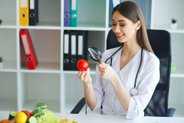 Health. The doctor signs out a diet plan. The Dietitian Holds in the Handfuls of Fresh Tomato. Fruits and Vegetables. Young Doctor with a Beautiful Smile at the Office. High Resolution