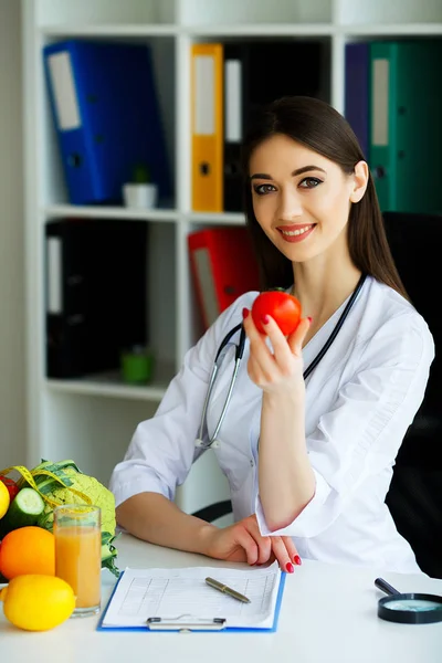 Health. The doctor signs out a diet plan. The Dietitian Holds in the Handfuls of Fresh Tomato. Fruits and Vegetables. Young Doctor with a Beautiful Smile at the Office. High Resolution