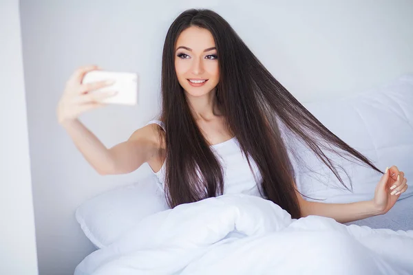 幸せなかわいい女のショットが携帯電話を使用してベッドの上にあります — ストック写真