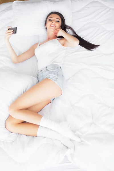 幸せなかわいい女のショットが携帯電話を使用してベッドの上にあります — ストック写真