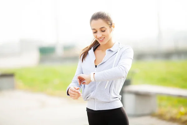 Foto de Joyful Fitness Woman 30s en ropa deportiva tocando el auricular Bluetooth y sosteniendo el teléfono móvil, mientras descansa en Green Park — Foto de Stock