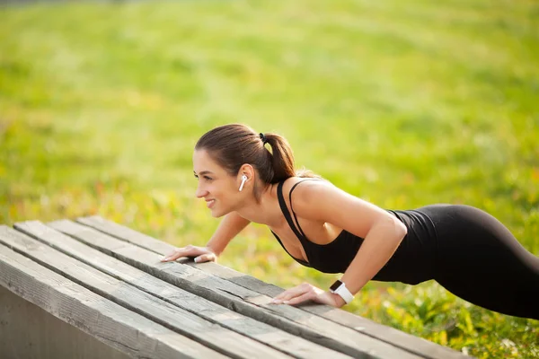 Portrait de belle femme sportive 20s en vêtements de sport faisant push-ups, et écouter de la musique avec écouteur bluetooth pendant l'entraînement dans un parc vert — Photo