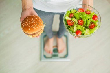 Diyet. Kadın vücut ağırlığı Holding Burger ve salata ölçek ağırlığında üzerinde ölçüm. Tatlılar sağlıksız abur cubur var. Diyet, sağlıklı beslenme, yaşam tarzı. Kilo kaybı. Obezite. Üstten görünüm