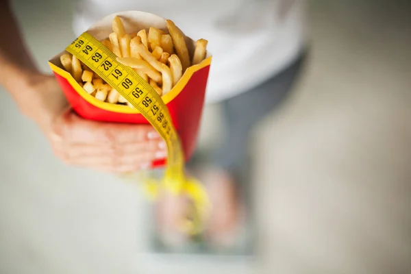 Диета. Женщина измеряет вес тела по весу, держа нездоровую вредную пищу. Потеря веса. Ожирение. Вид сверху — стоковое фото