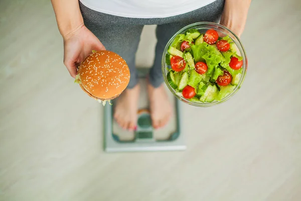 Дієта. Жінка вимірює вагу тіла на ваговій шкалі утримуючи бургер та салат. Солодощі є нездоровою їжею. Дієта, здорове харчування, спосіб життя. Втрата ваги. Ожиріння. Вид зверху — стокове фото