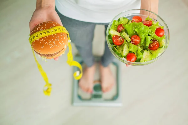 Dieta. Mujer que mide el peso corporal en la balanza que sostiene la hamburguesa y la ensalada. Los dulces son comida chatarra malsana. Dieta, Comida Saludable, Estilo de Vida. La pérdida de peso. Obesidad. Vista superior — Foto de Stock