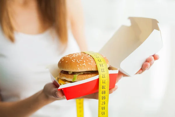 Dieta. Una joven sosteniendo una hamburguesa en una cinta métrica. El concepto de alimentación saludable — Foto de Stock