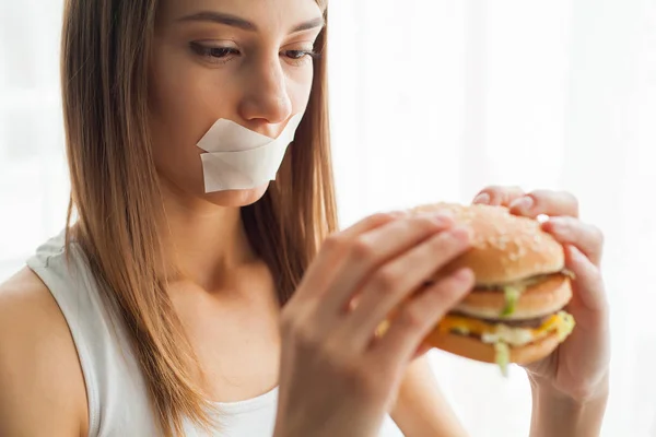 Diet. Young woman with duct tape over her mouth, preventing her to eat junk food. Healthy eating concept
