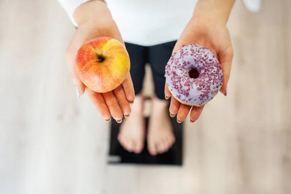 Dieta. Mujer que mide el peso corporal en la balanza de pesaje que sostiene la rosquilla y la manzana. Los dulces son comida chatarra malsana. Dieta, Comida Saludable, Estilo de Vida. La pérdida de peso. Obesidad —  Fotos de Stock