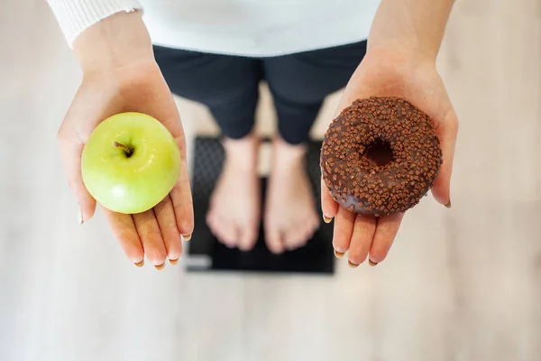 Δίαιτα. Γυναίκα μέτρησης σώματος βάρος για ζύγιση κλίμακας κρατώντας ντόνατ και μήλο. Τα γλυκά είναι ανθυγιεινά τρόφιμα παλιοπραγμάτων. Να κάνει δίαιτα, υγιεινή διατροφή, τον τρόπο ζωής. Απώλεια βάρους. Η παχυσαρκία — Φωτογραφία Αρχείου