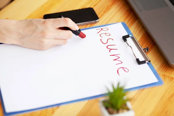 Búsqueda de trabajo. La mano femenina escribe curriculum vitae con lápiz labial en una hoja de papel blanco. Escritorio de oficina de madera con ordenador portátil, smartphone y suministros — Foto de Stock