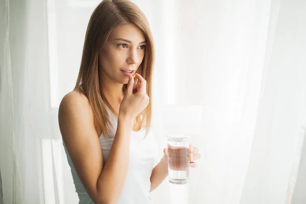 Mutlu gülümseyen pozitif kadın hap yiyor ve evinde elinde bir bardak su tutuyor.