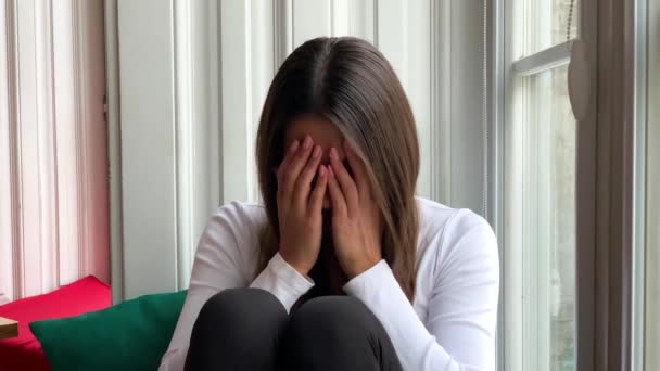 Mujer joven y triste llorando sentada en una ventana en casa — Vídeos de Stock