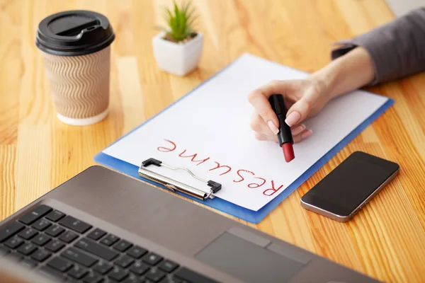 Job search. Female hand writes resume with lipstick on white sheet of paper. Wooden office desk with laptop, smartphone and supplies — Stock Photo, Image