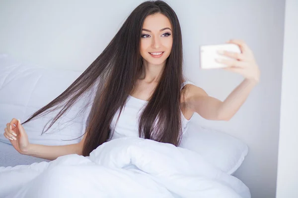 幸せなかわいい女のショットが携帯電話を使用してベッドの上にあります。 — ストック写真