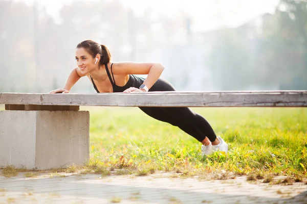 Πορτρέτο του όμορφη γυναίκα σπορ 20s σε αθλητικά είδη κάνει push-ups, και ακούγοντας μουσική με το κάλυμμα ακουστικού bluetooth κατά τη διάρκεια της προπόνηση σε καταπράσινο πάρκο — Φωτογραφία Αρχείου