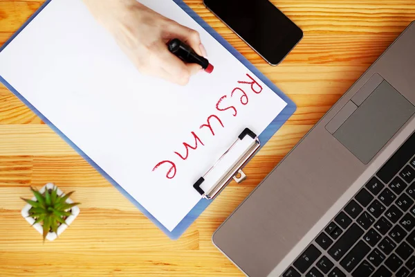 Job search. Female hand writes resume with lipstick on white sheet of paper. Wooden office desk with laptop, smartphone and supplies — Stock Photo, Image