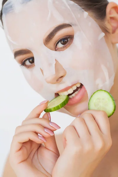 Beautiful young woman is getting facial clay mask at spa, lying with cucumbers on eyes — Stock Photo, Image