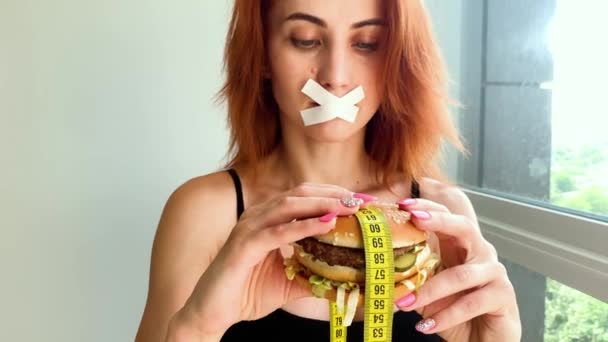 Kost. Porträtt av en kvinna vill äta en hamburgare, men en limmad mun, en uppfattning om kost, ohälsosam mat, en vilja i näring — Stockvideo