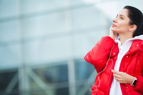 Foto einer fröhlichen Fitness-Frau in Sportbekleidung, die Bluetooth-Ohrhörer berührt und Handy hält, während sie sich im grünen Park ausruht — Stockfoto