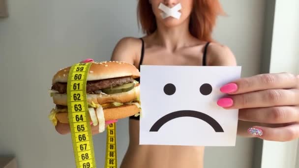 Diyet. Bir kadının portresi bir burger yemek istiyor, ama bir yapıştırılmış ağız, diyet kavramı, sağlıksız gıda, beslenme bir irade — Stok video