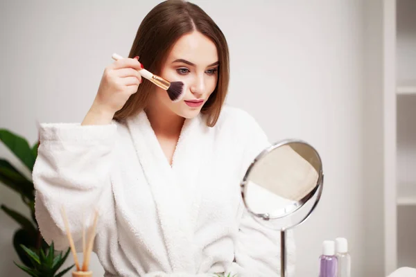 Mujer joven en casa se aplica maquillaje en la cara en el baño delante de un espejo — Foto de Stock