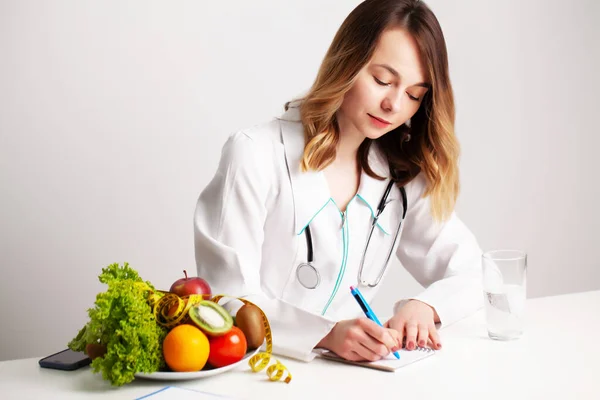 Médico dietista joven en la sala de consulta en la mesa con verduras y frutas frescas, trabajando en un plan de dieta —  Fotos de Stock