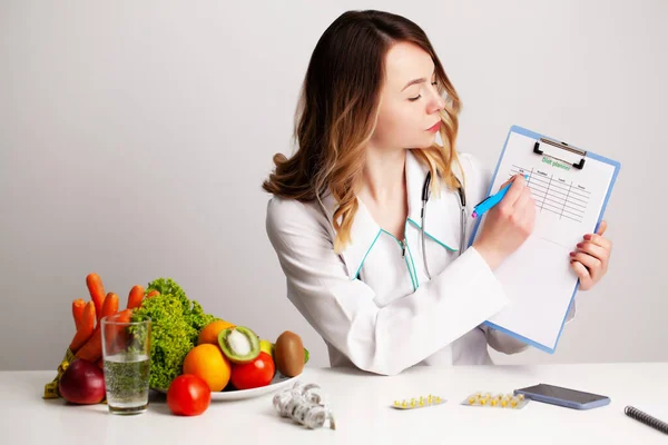 Médico dietista joven en la sala de consulta en la mesa con verduras y frutas frescas, trabajando en un plan de dieta — Foto de Stock