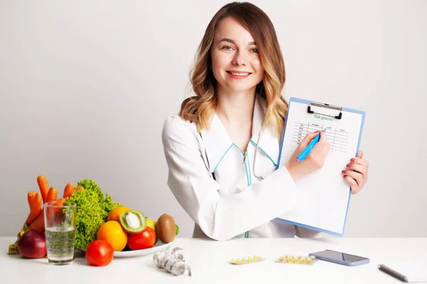 Médico dietista joven en la sala de consulta en la mesa con verduras y frutas frescas, trabajando en un plan de dieta — Foto de Stock