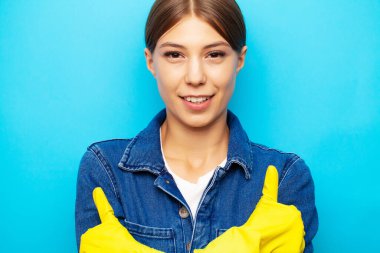 Önlüklü ve sarı eldivenli bir temizlik şirketinin çalışanı mavi arka planda poz veriyor.